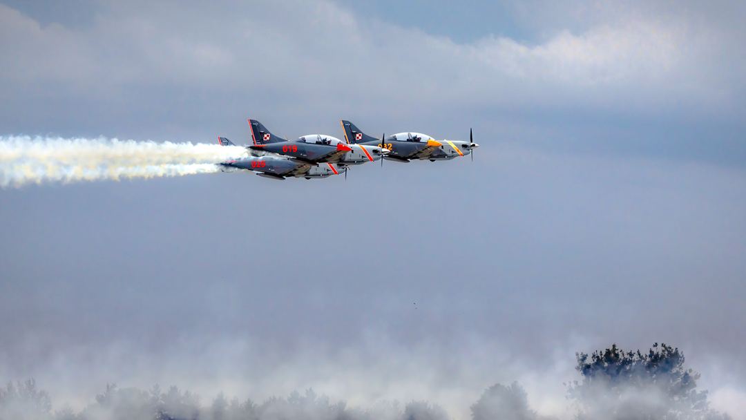 In aviation, weather isn't merely a backdrop—it dictates the entire show. Instead of clear blue skies, the frame is shaped by a heavy, overcast ceiling and misty haze hugging the treeline, forcing the team into a low-altitude "flat display" with no vertical maneuvers.
The creative approach was to embrace this meteorological moodiness rather than fight it. The dense, saturated air catches and holds the thick smoke trails, while the tight formation of PZL-130 Orlik turboprops slicing across the horizon creates a sharp contrast between a somber front and cold mechanical precision. Rather than compromising the shot, adverse weather provided a dramatic, natural stage—highlighting the sheer discipline of pilots operating right below the cloud base.
