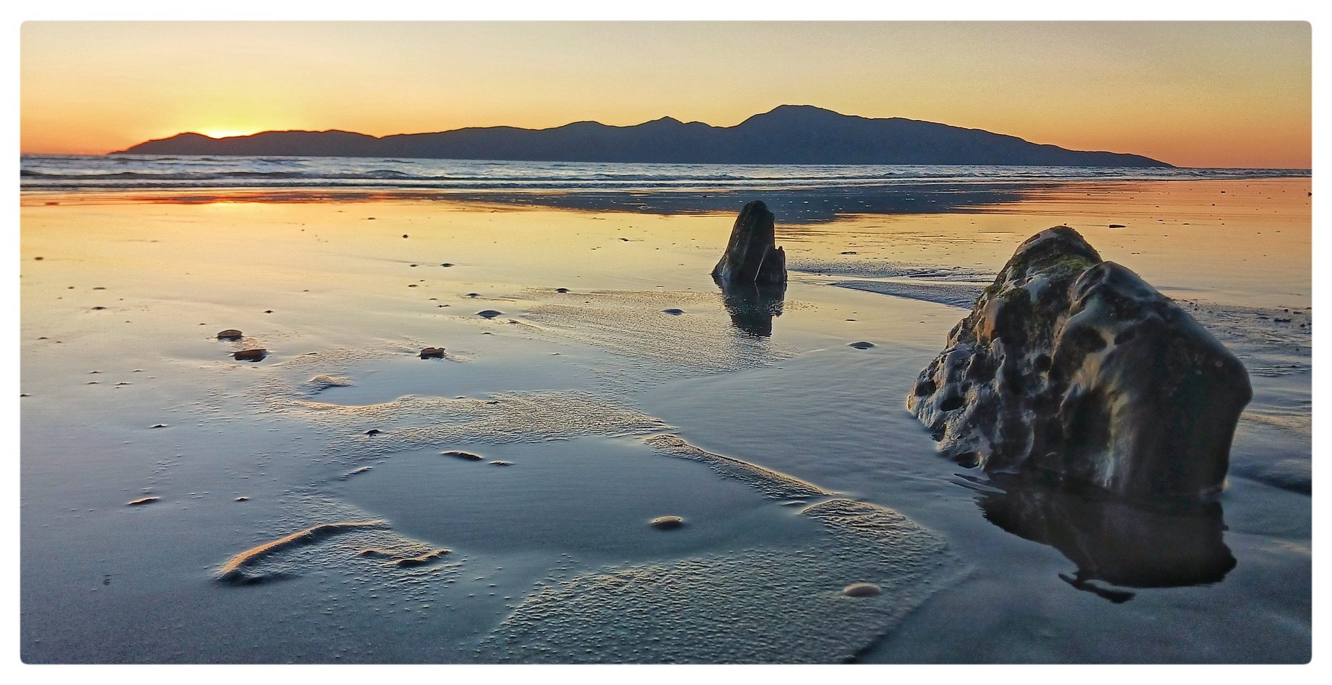 Kāpiti Island on the horizon at sunset, with wet sand, pools and dark, mostly buried driftwood in the foreground.