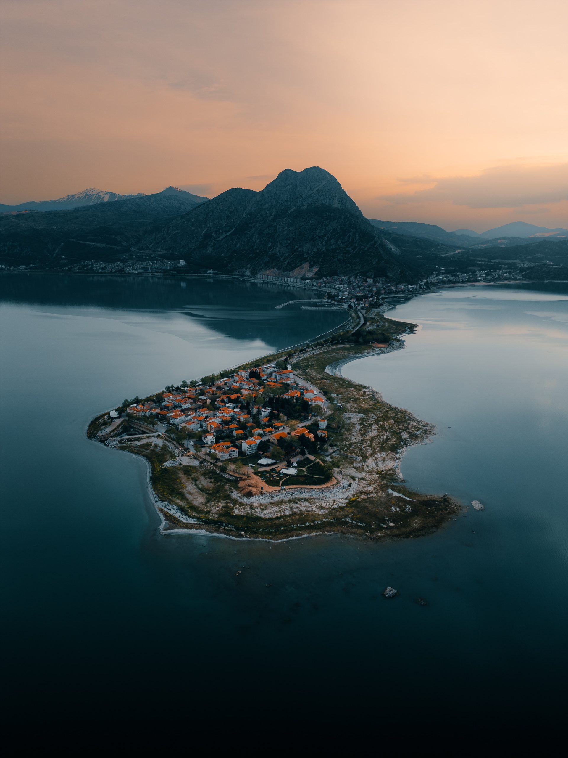 An aerial view at dusk of a village on a narrow peninsula in a lake, with mountains behind