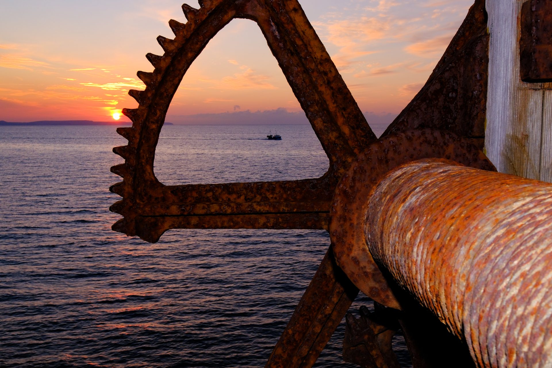 The sun rising over the sea just beside the teeth of a large rusty gear wheel, with a small boat on the water framed inside the wheel and a rusted cable drum in the foreground.