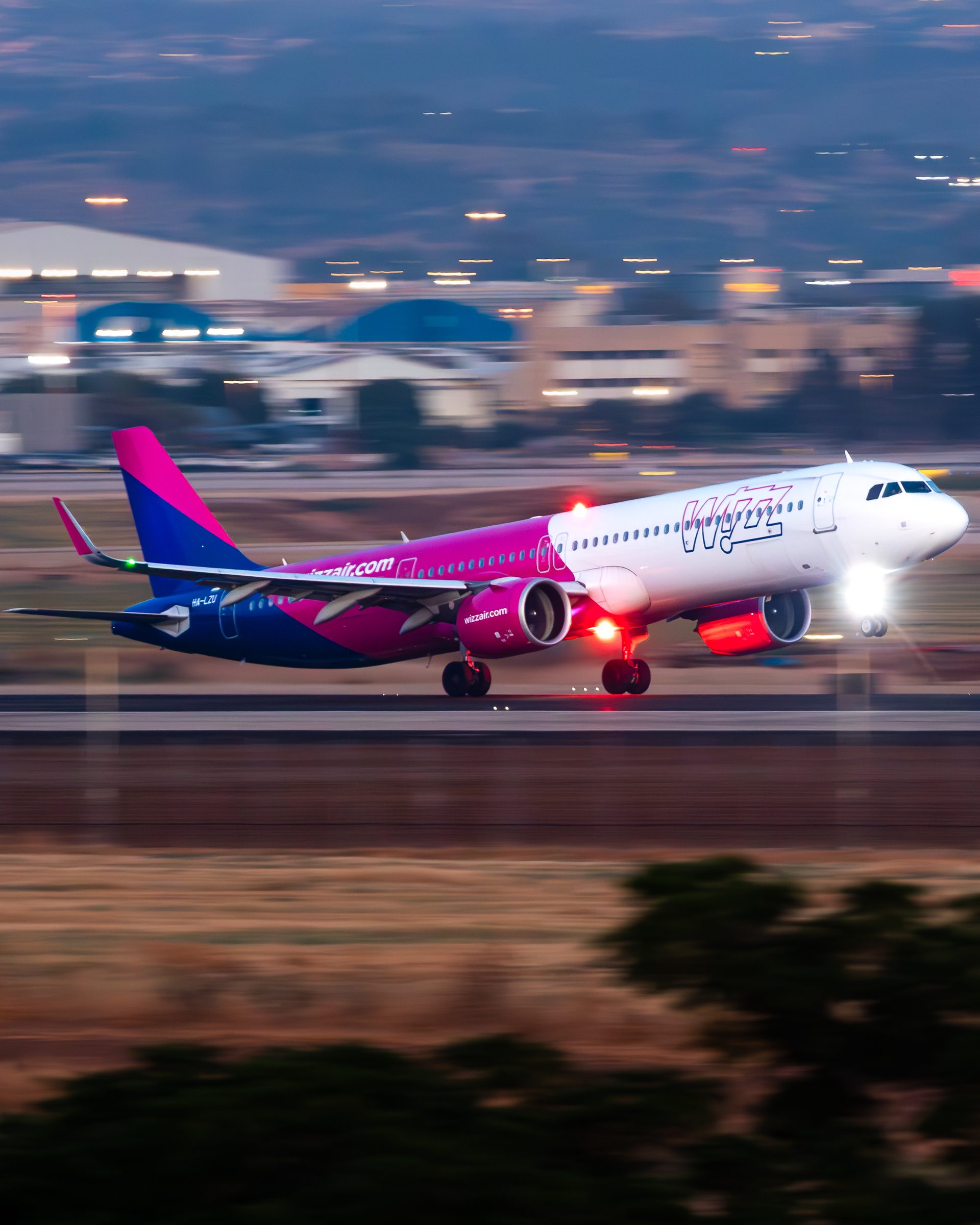 A Wizz Air airliner lifting its nose wheel on take-off at dusk with its landing lights on, and blurred airport buildings and lit blue hills behind.