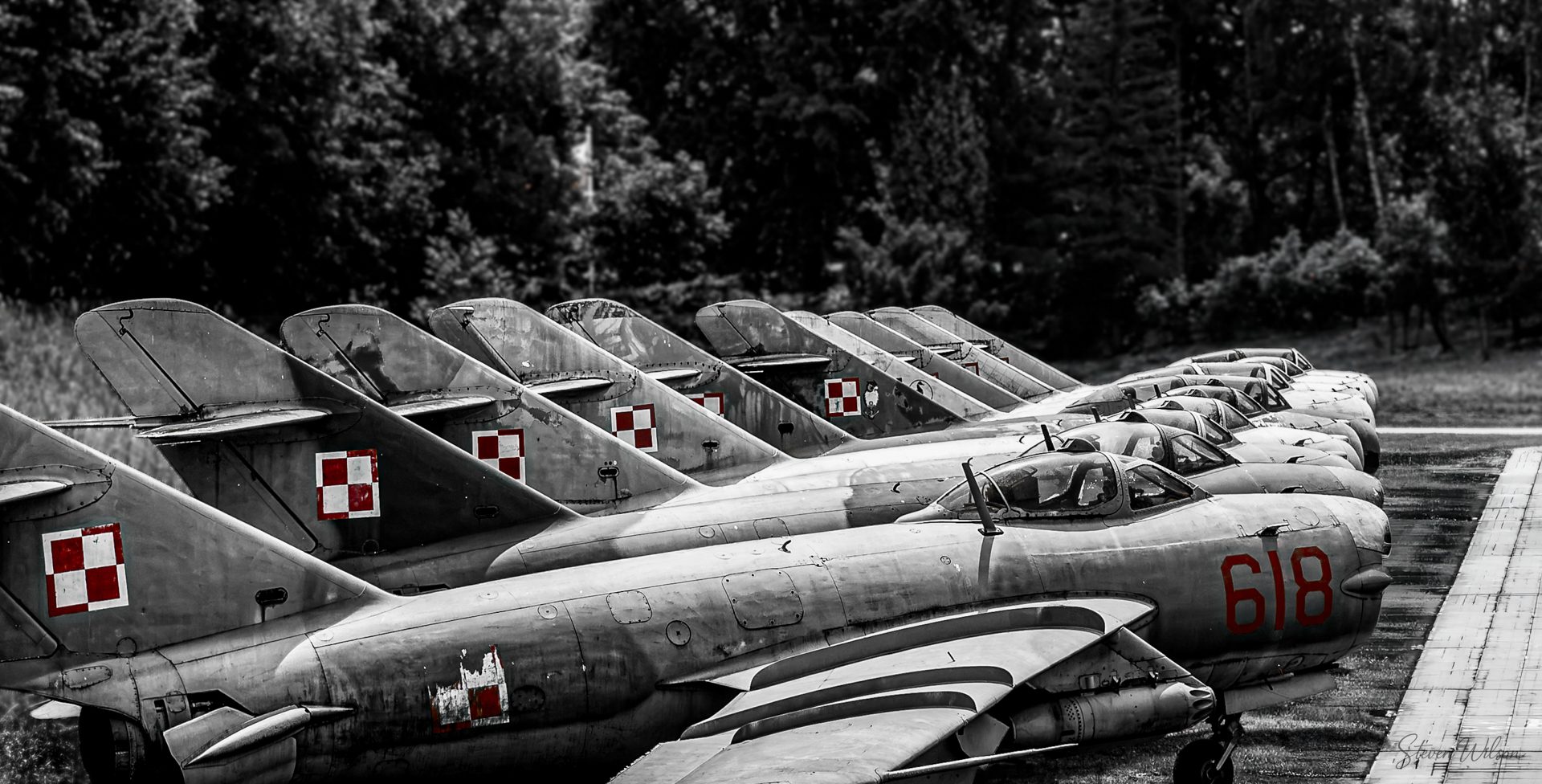 A line of Cold War jets parked at a museum, in black and white with the red and white checkerboard markings left in colour.