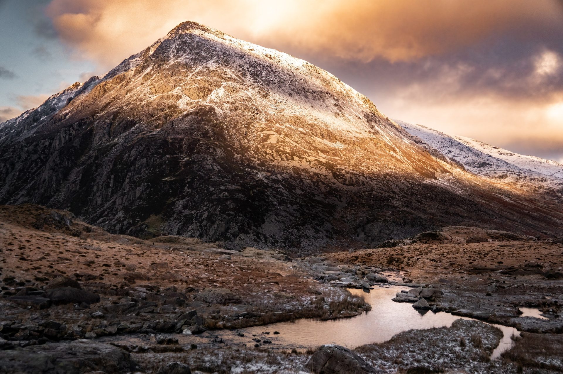 A snow-dusted mountain peak lit from the side by low sun under orange cloud, with a stream winding through rocky ground in front.