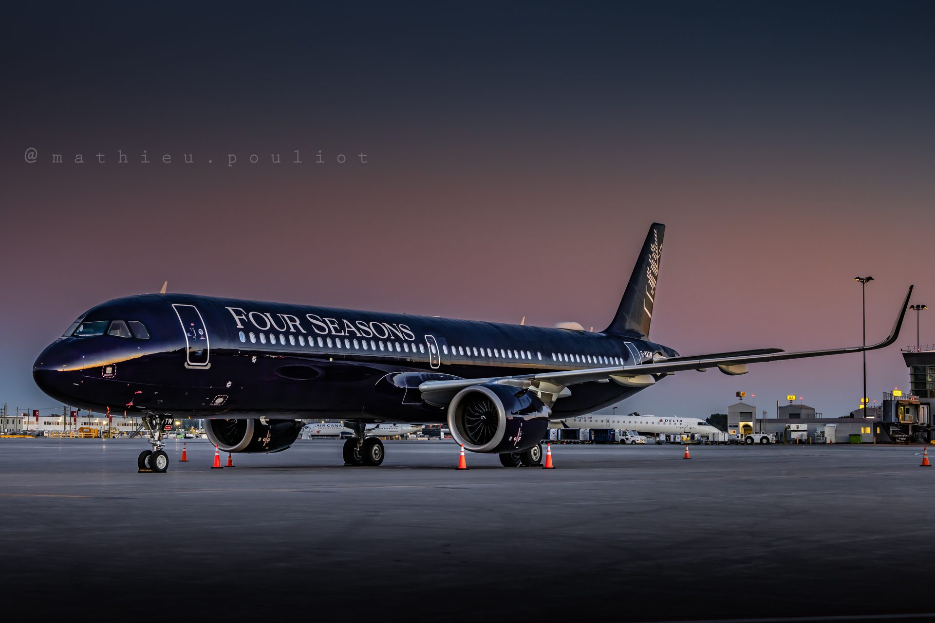 A dark Four Seasons A321 parked on an airport ramp before sunrise, with traffic cones around it and a pink and blue sky behind.