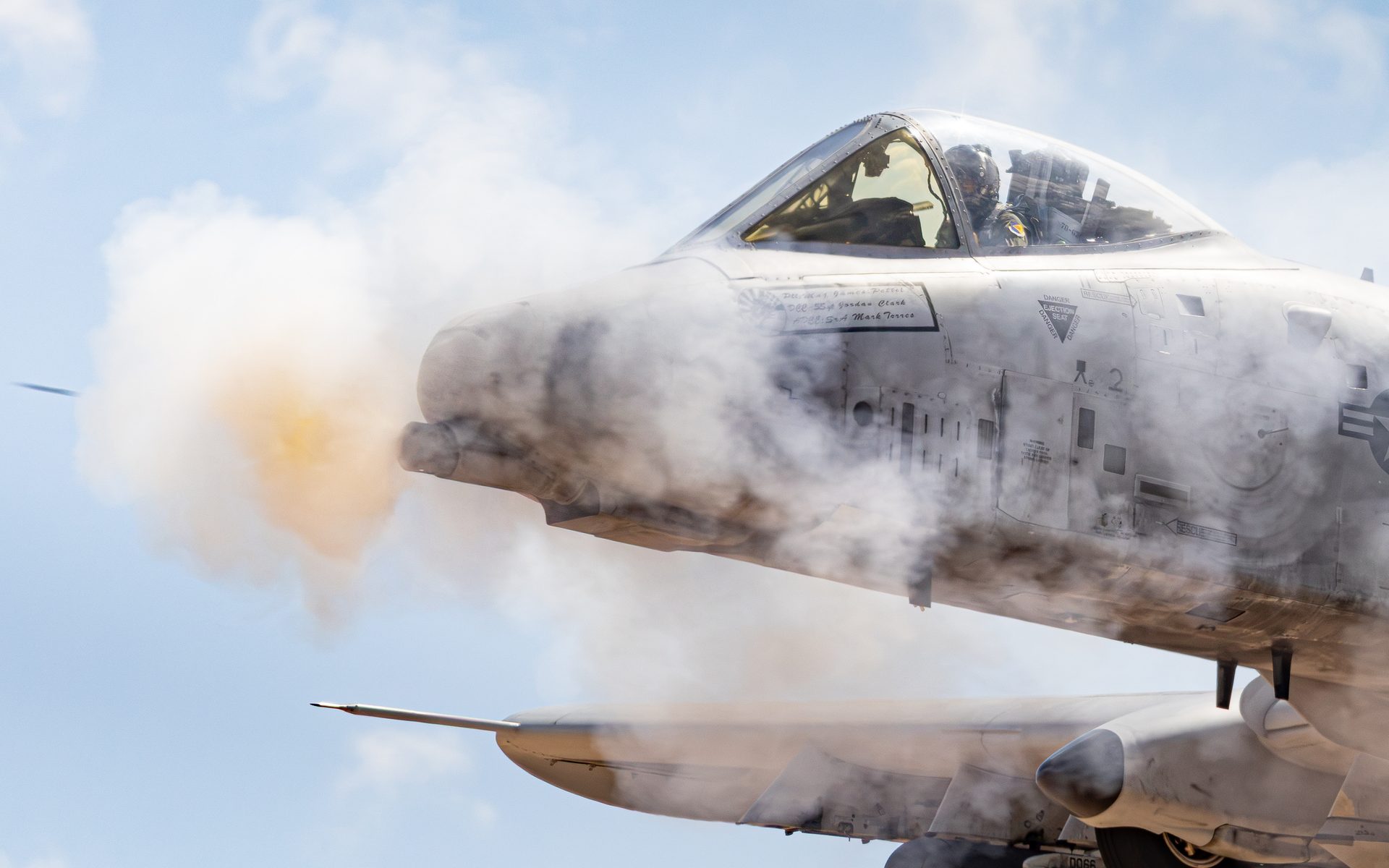 A close view of an A-10 firing its cannon, gun smoke rolling back over the sunlit nose and the pilot visible in the cockpit.