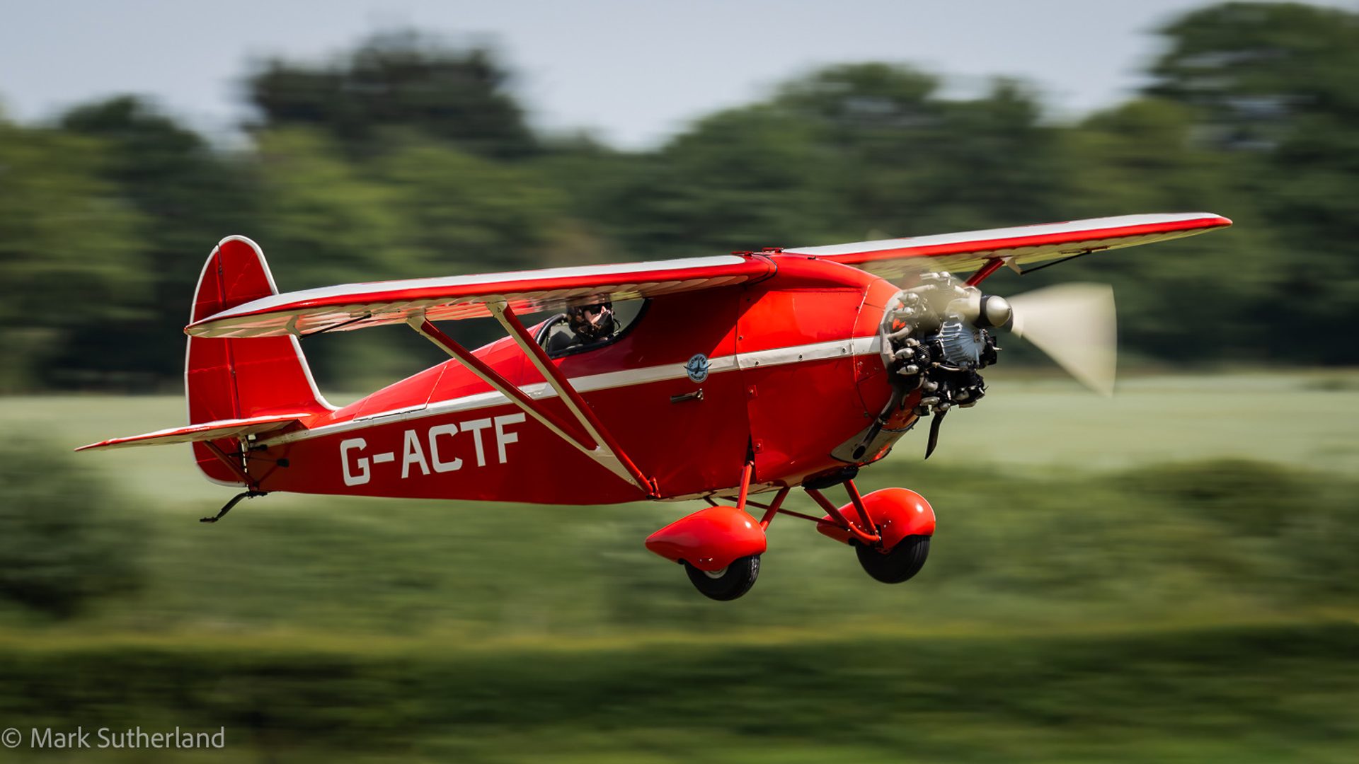 A red Comper Swift flying low, panned so the aircraft is sharp, the propeller a blur and the trees behind pulled into streaks.