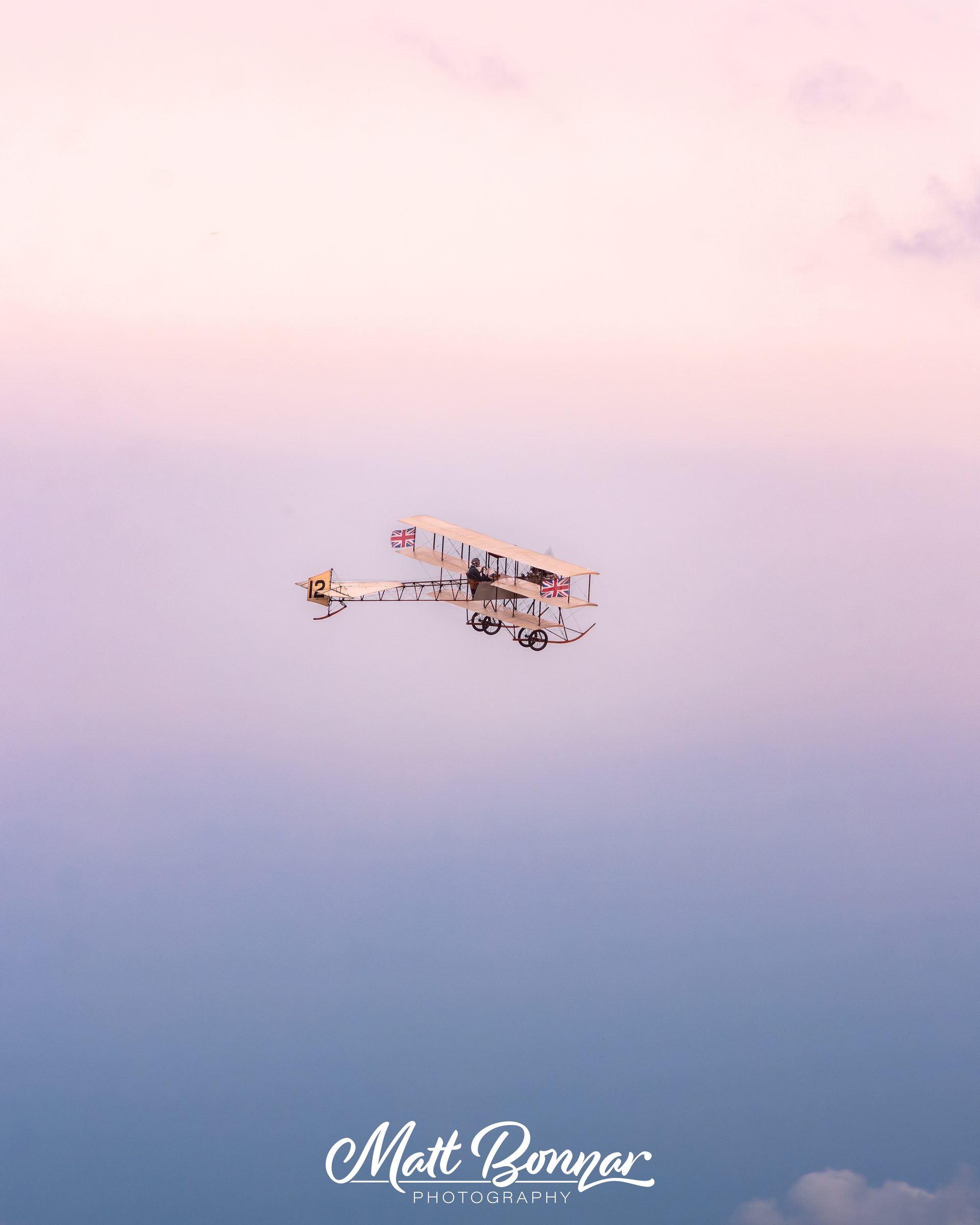 The Avro Triplane, an early wood-and-wire aircraft with Union flags on it, flying small in the middle of a soft pink and blue evening sky.