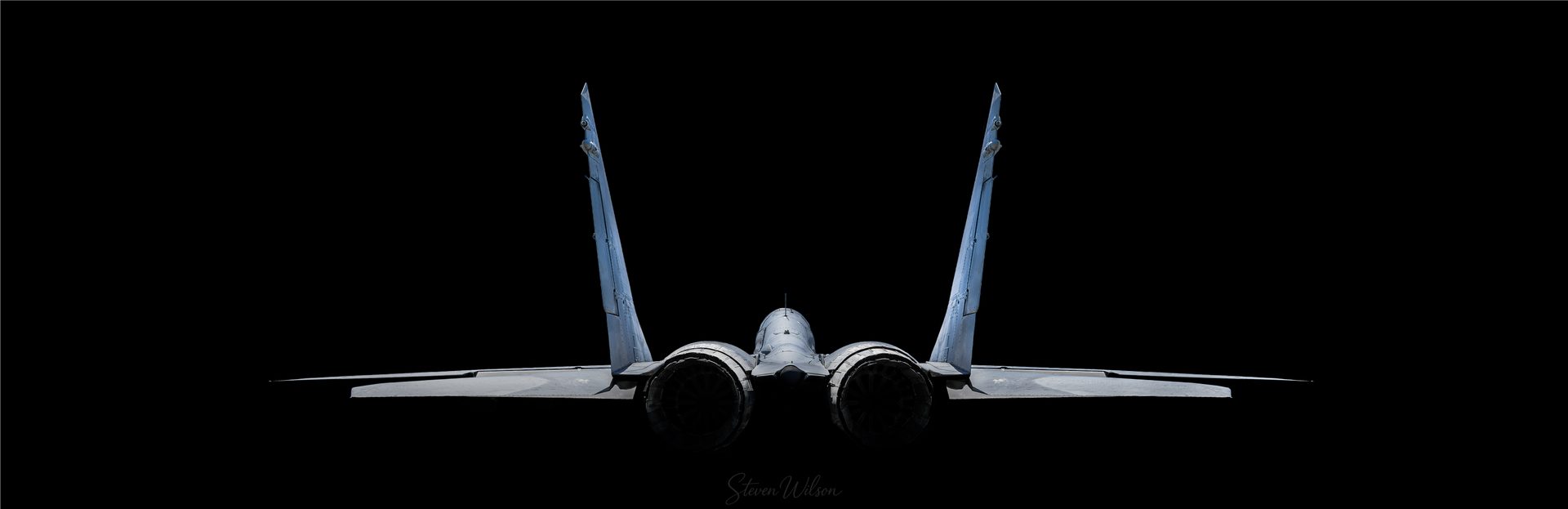The rear of a MiG-29 in a low key edit, the jet lit against a completely black background.