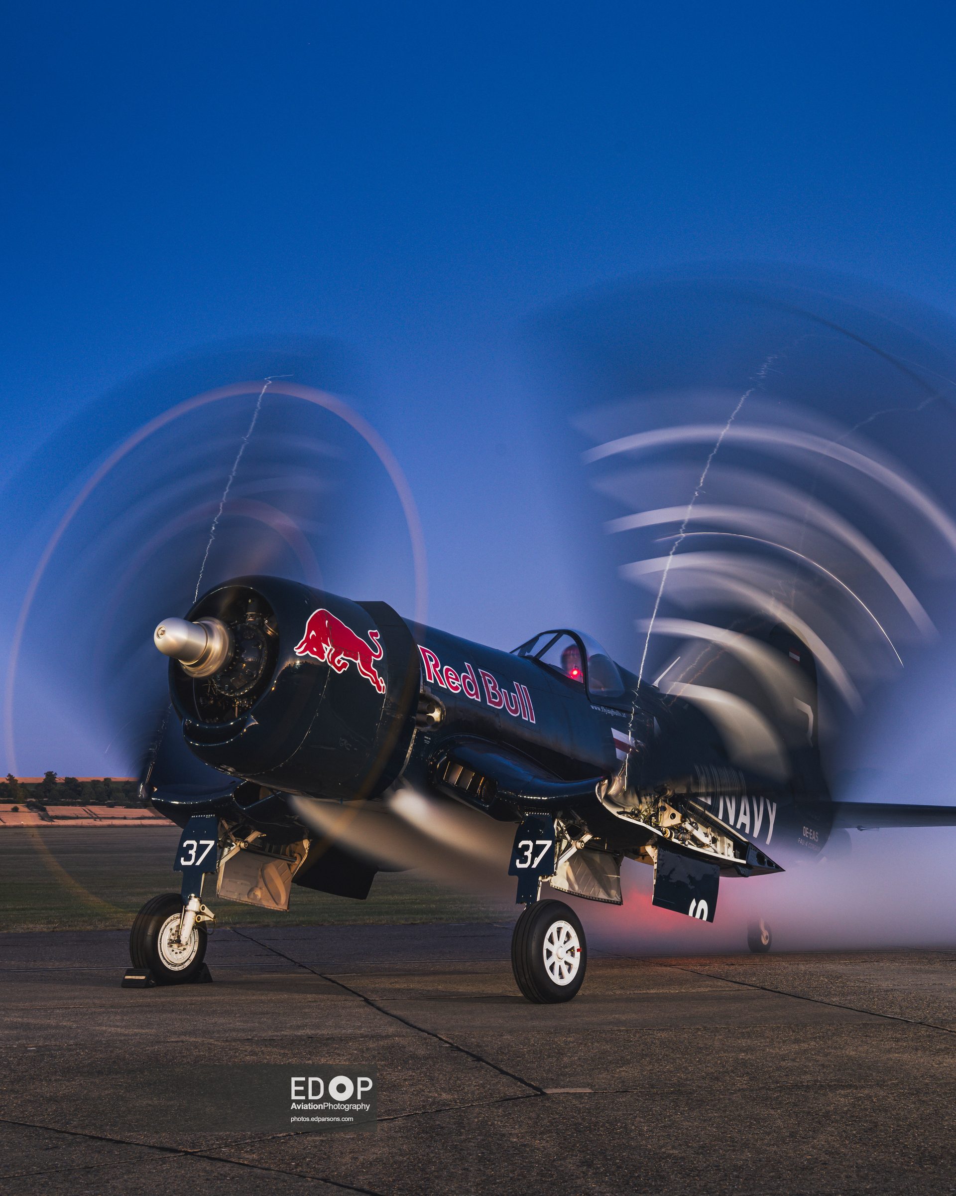 A Red Bull Corsair on the apron at dusk against a deep blue sky, its turning propeller blurred into a swirl by a long exposure.