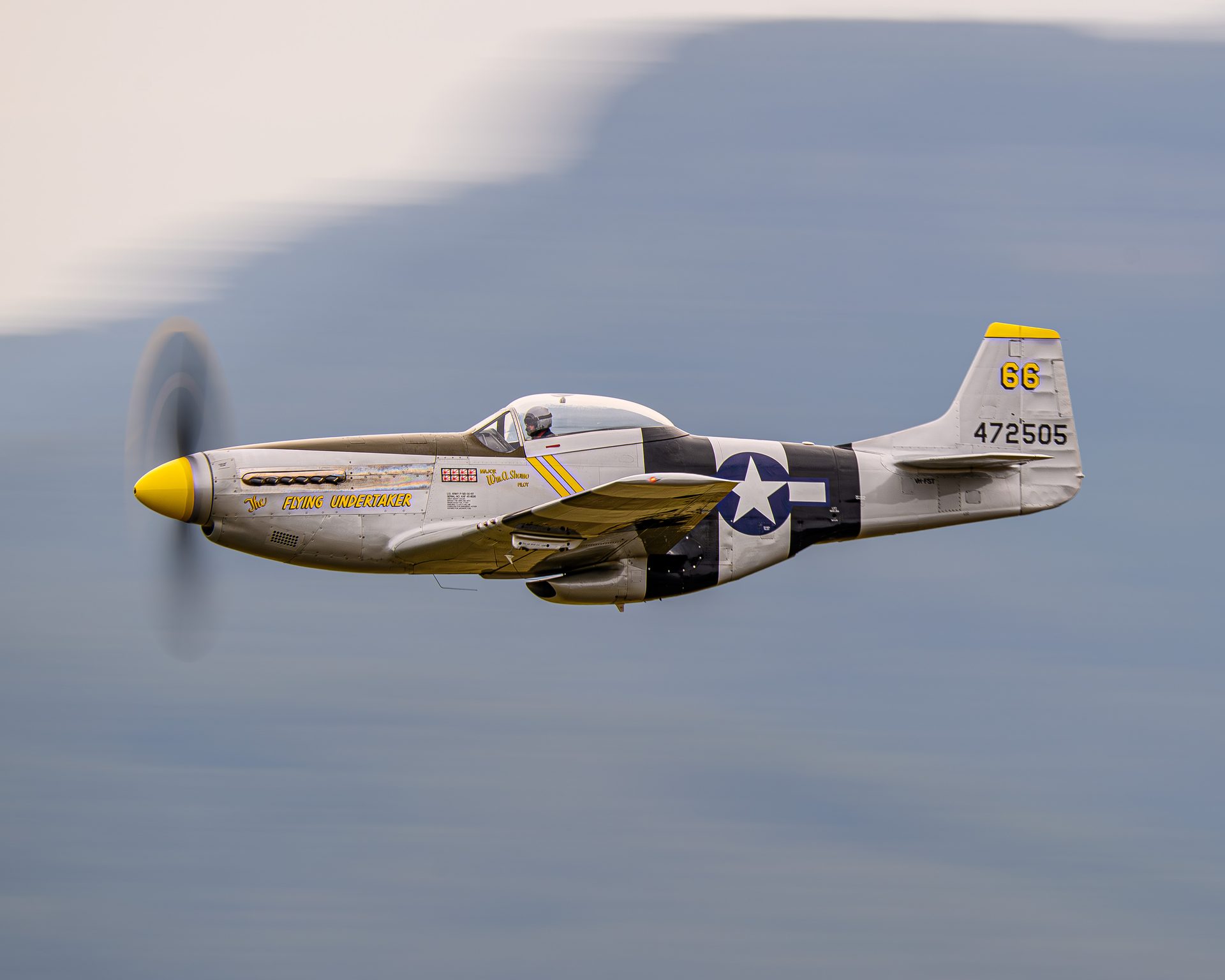 A silver P-51 Mustang with a yellow nose in flight, the propeller a blurred disc and the background smeared into streaks.