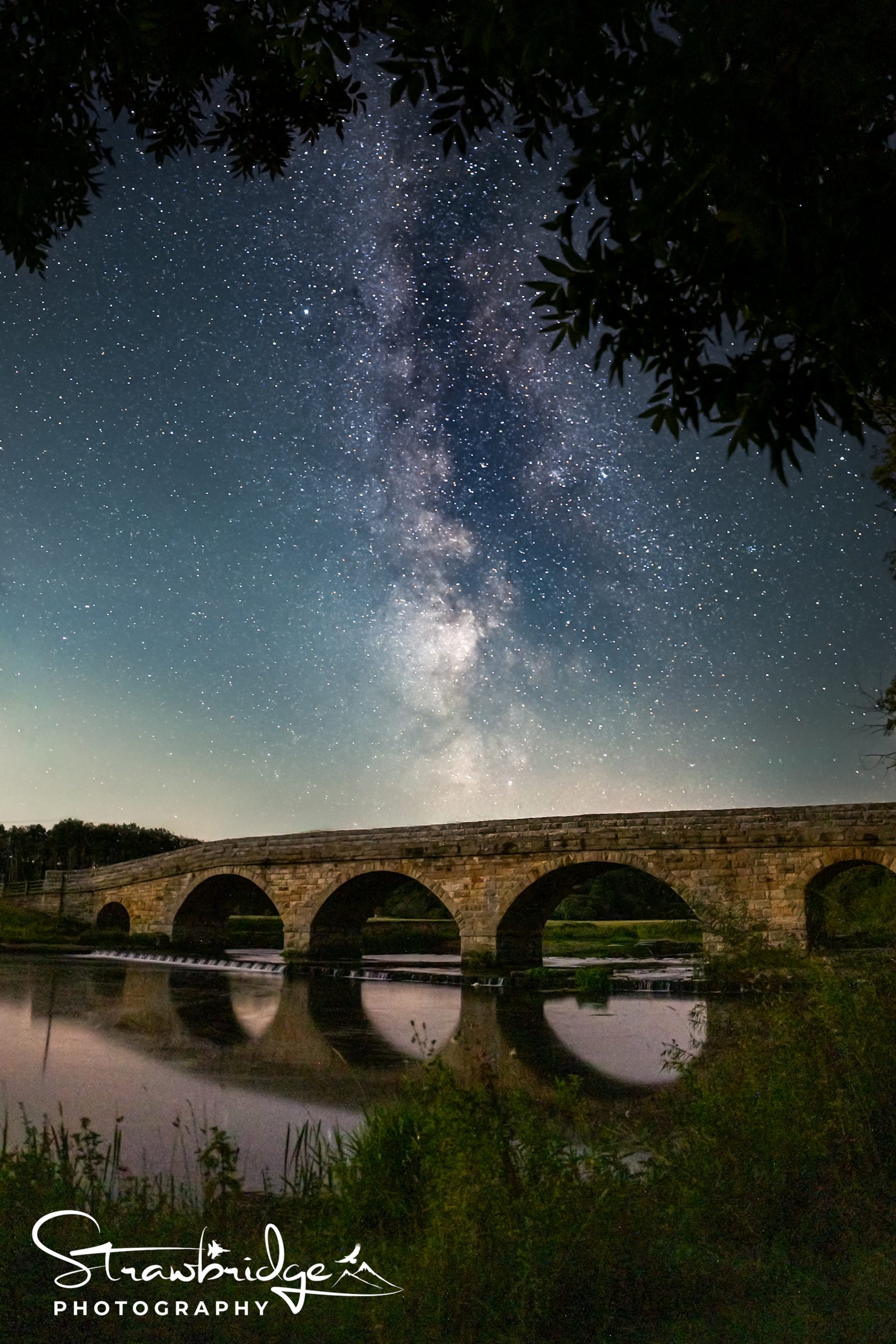 The Milky Way over a long stone bridge with arches, reflected in a river at night.