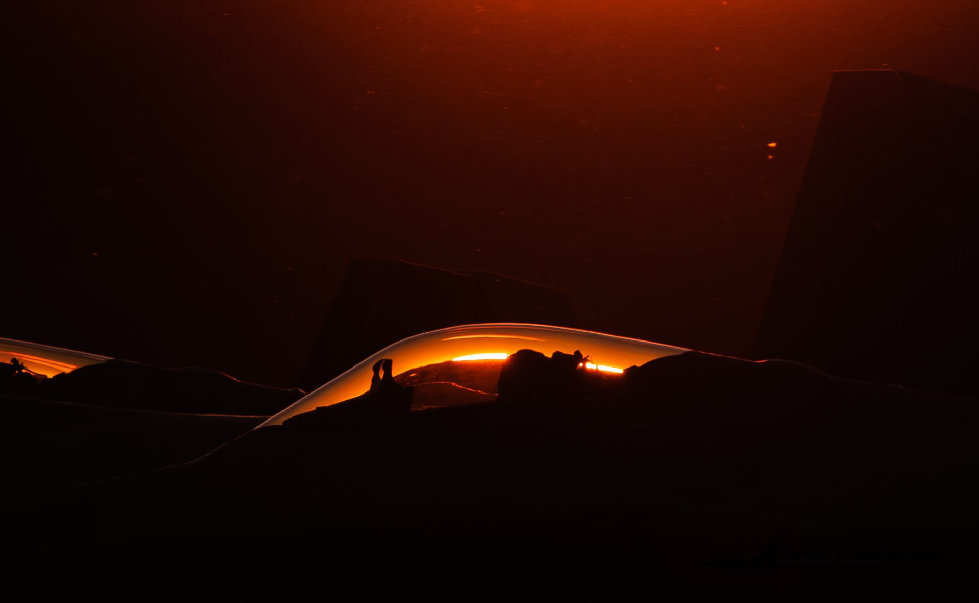 An F-22 Raptor almost in silhouette at sunset, with orange light glowing through the cockpit canopy