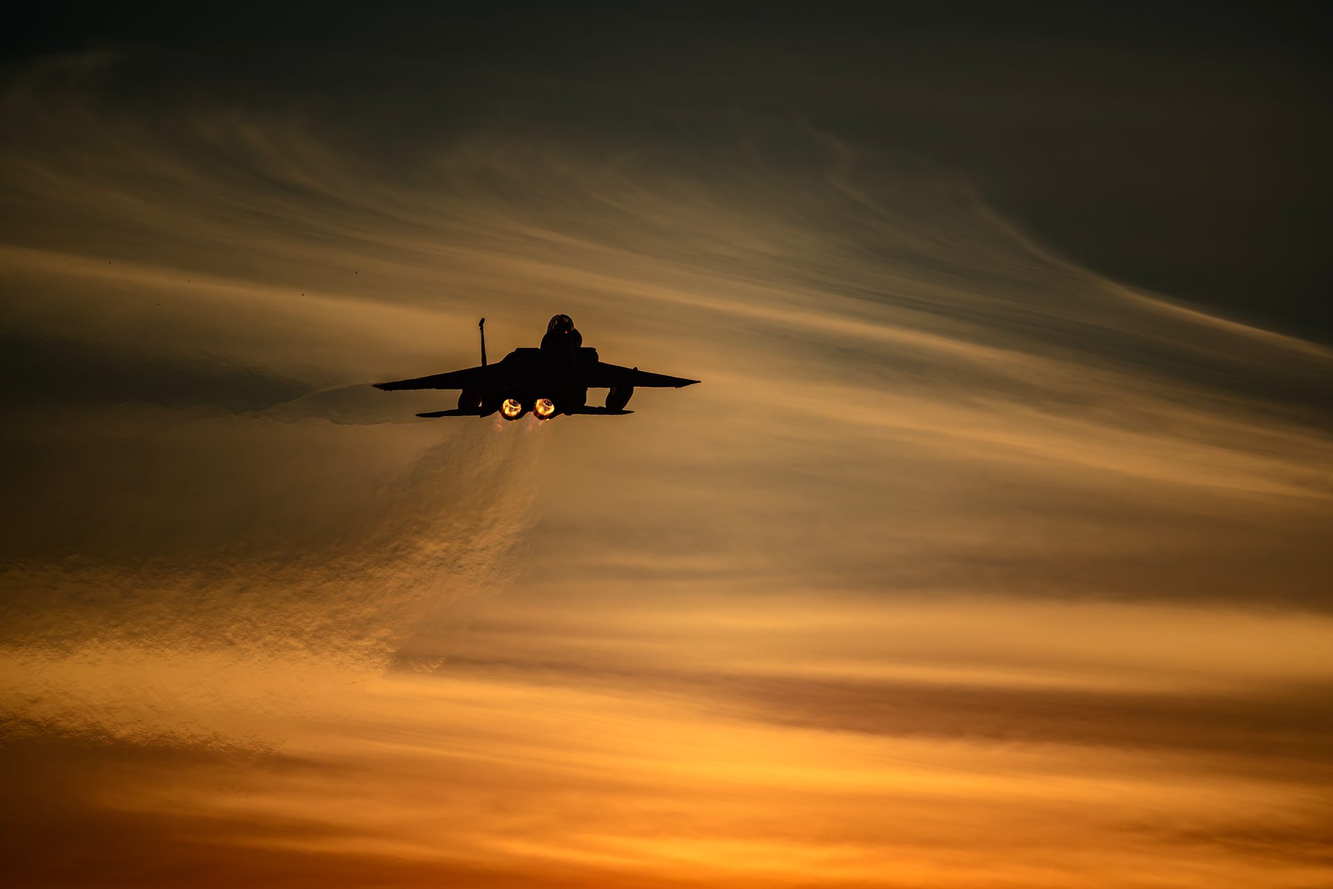 A fighter jet climbing away into an orange sunset sky, in silhouette with both afterburners glowing