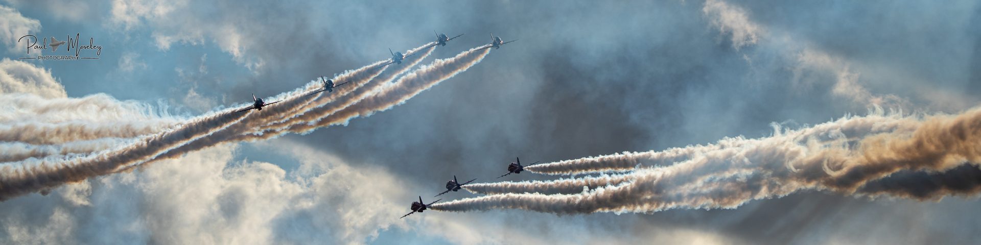 Two groups of Red Arrows jets crossing in opposite directions, trailing white smoke across a cloudy sky.