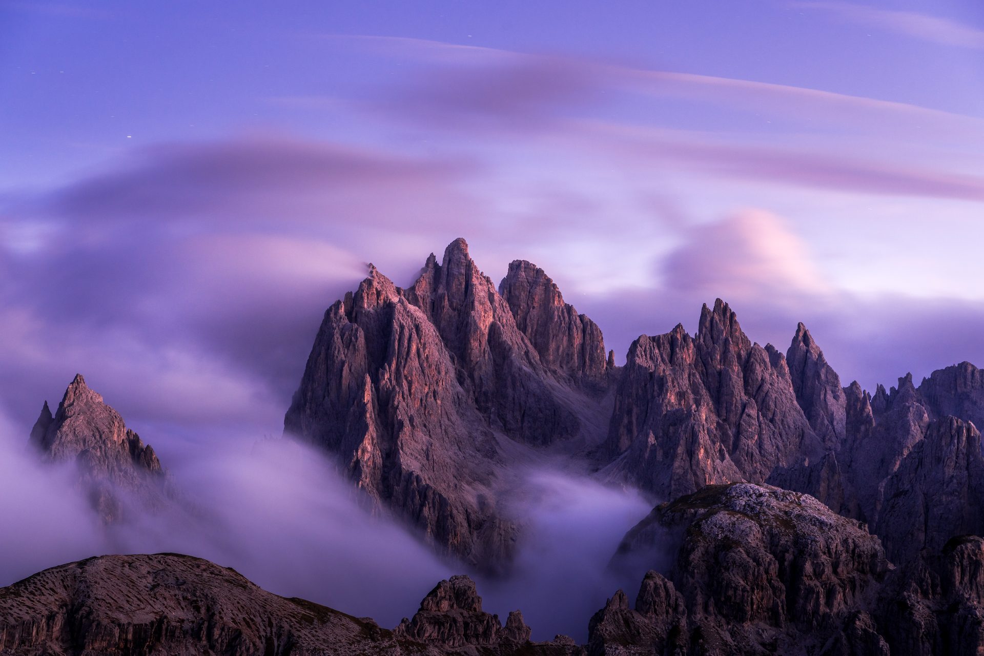 Jagged mountain peaks in the Dolomites under a purple and blue sky, with cloud smoothed by a long exposure drifting between them.