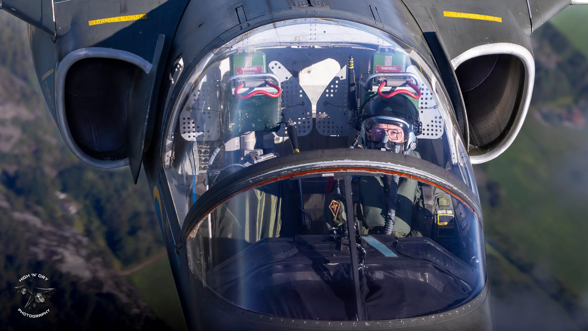 A tight crop of a SAAB SK-60 cockpit seen from above, with a crew member in helmet, glasses and oxygen mask looking up through the canopy.