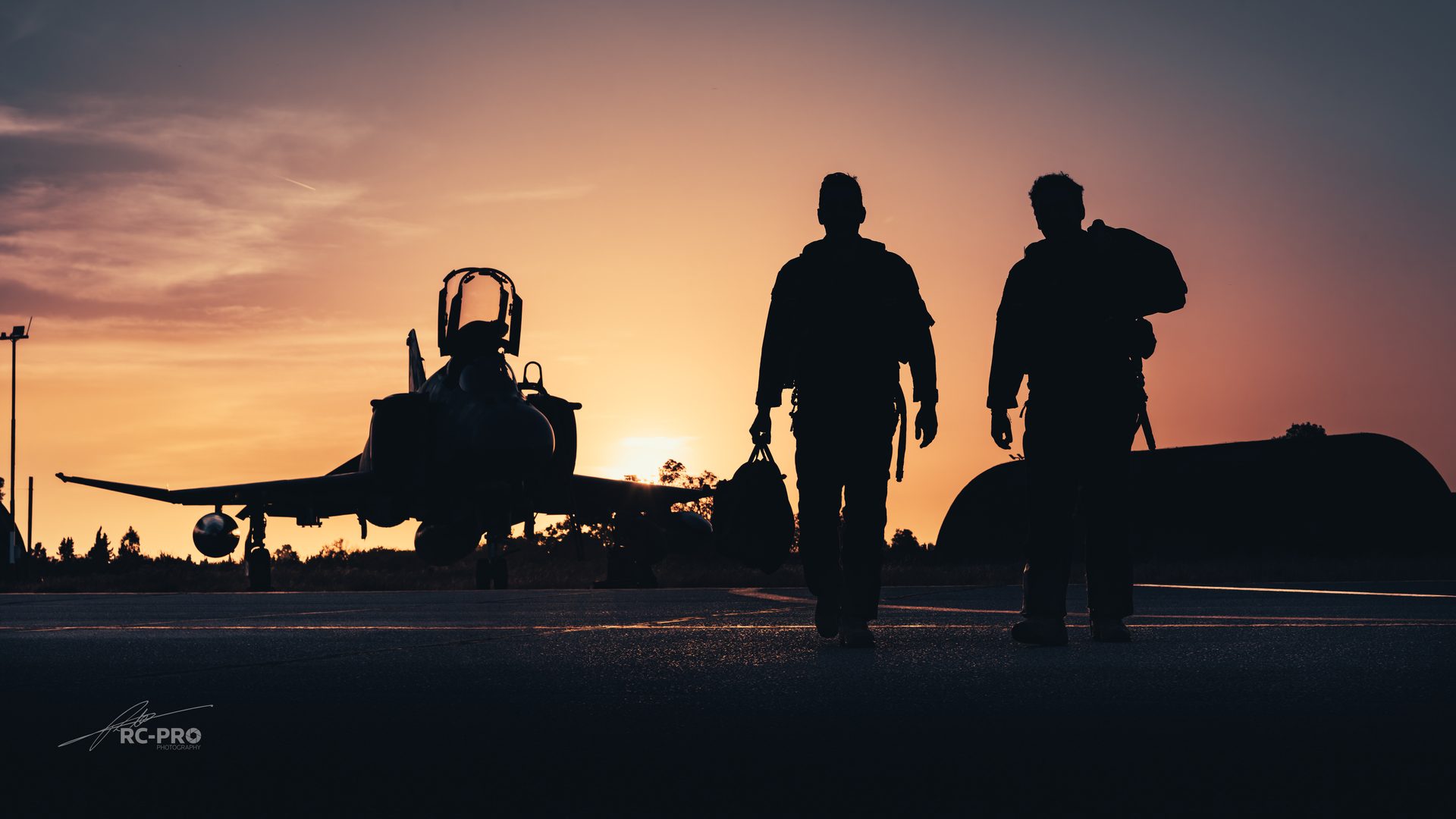 Two Phantom crew silhouetted against a sunset, walking across the apron with their kit, the jet parked behind them with its canopy open.