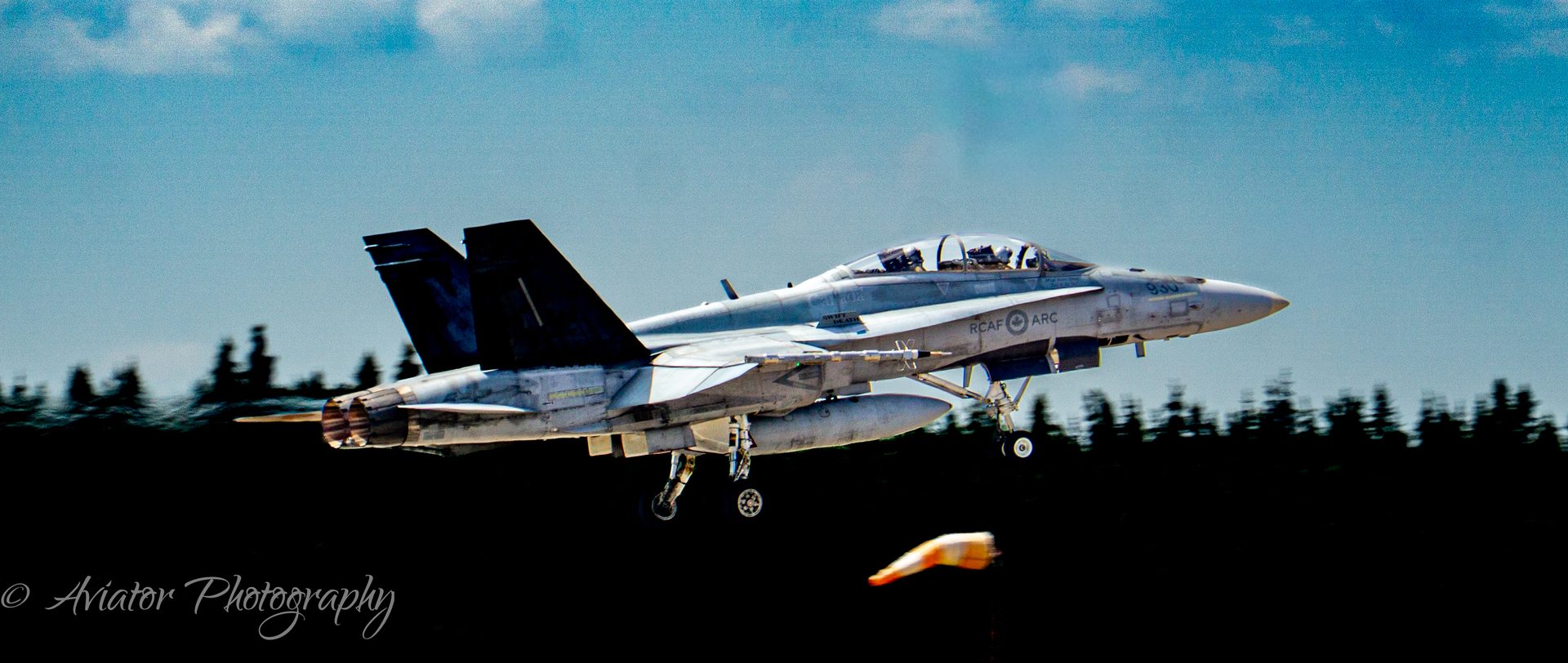 A CF-18 Hornet taking off with its gear down, above a dark treeline and an orange windsock.