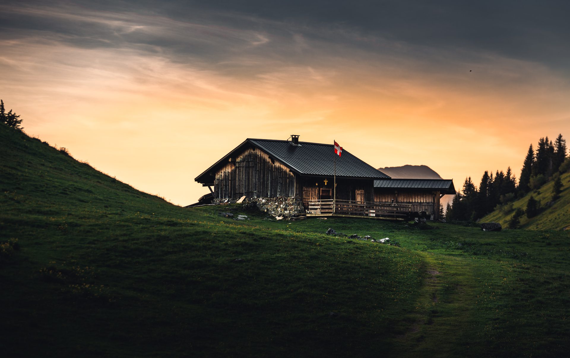 A wooden mountain hut with a flag on a grassy slope, dark against an orange sunset sky, with pine trees to the right.