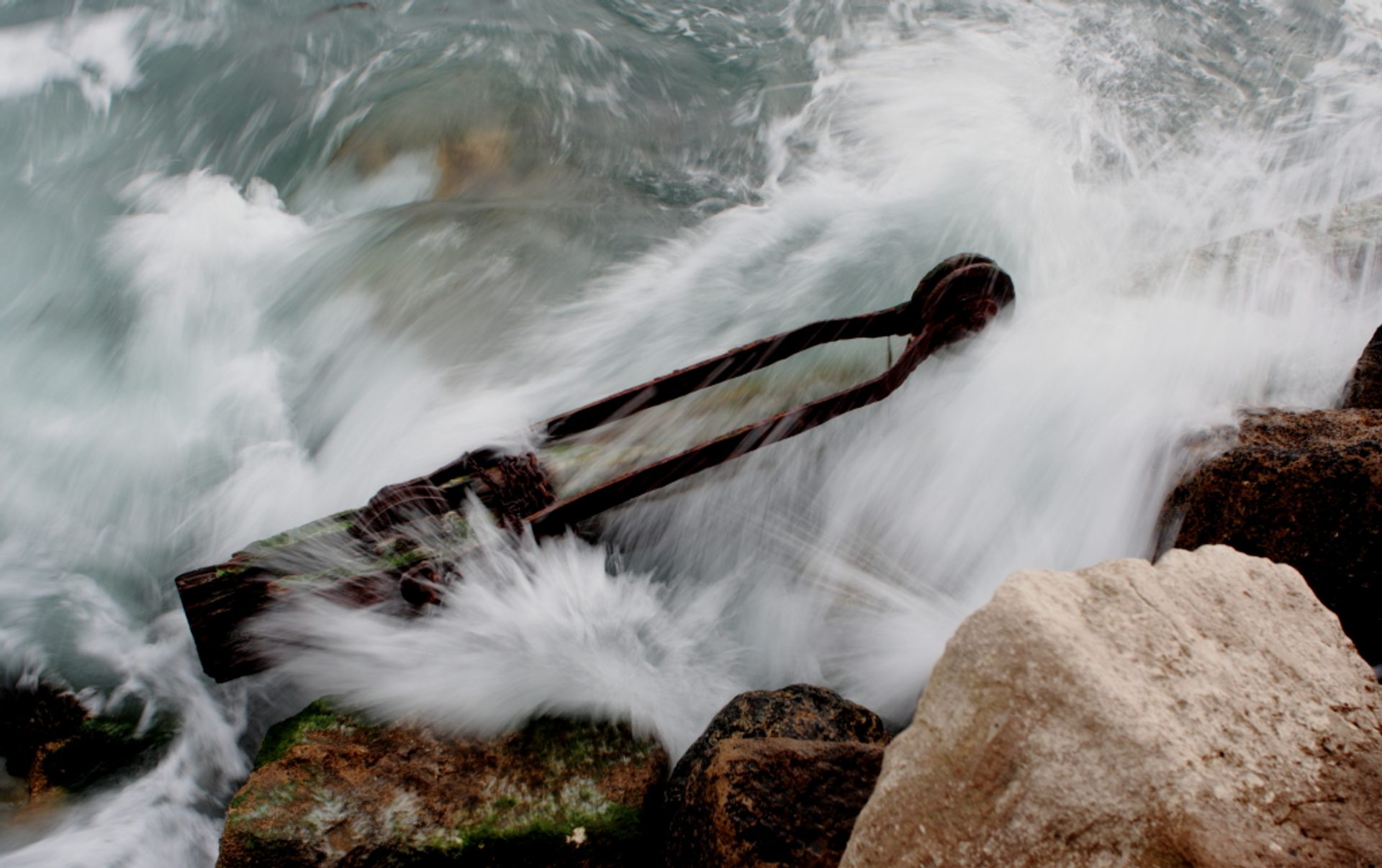 A wave breaking over a rusted piece of old crane lying in the sea between rocks, the spray blurred into white streaks.