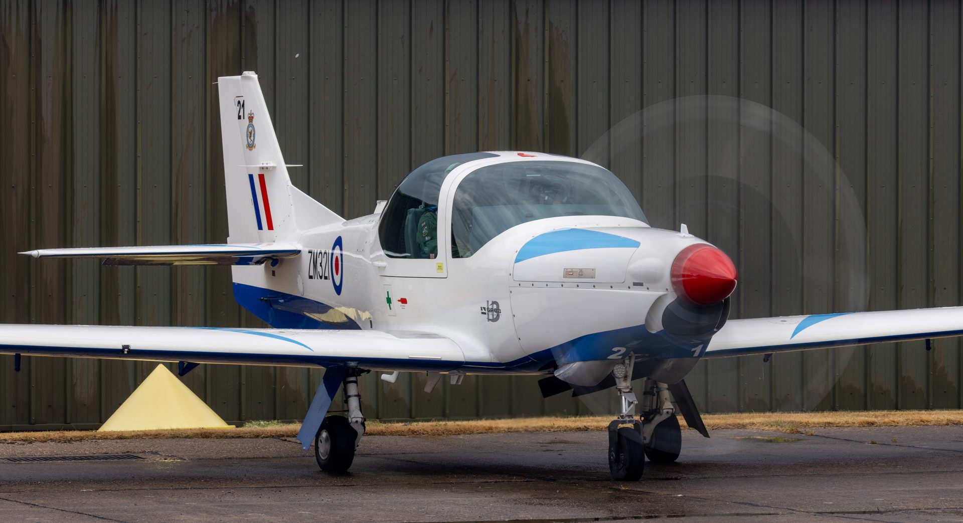 An RAF Prefect trainer on the ground in front of a hangar, its propeller blurred into a faint full disc.