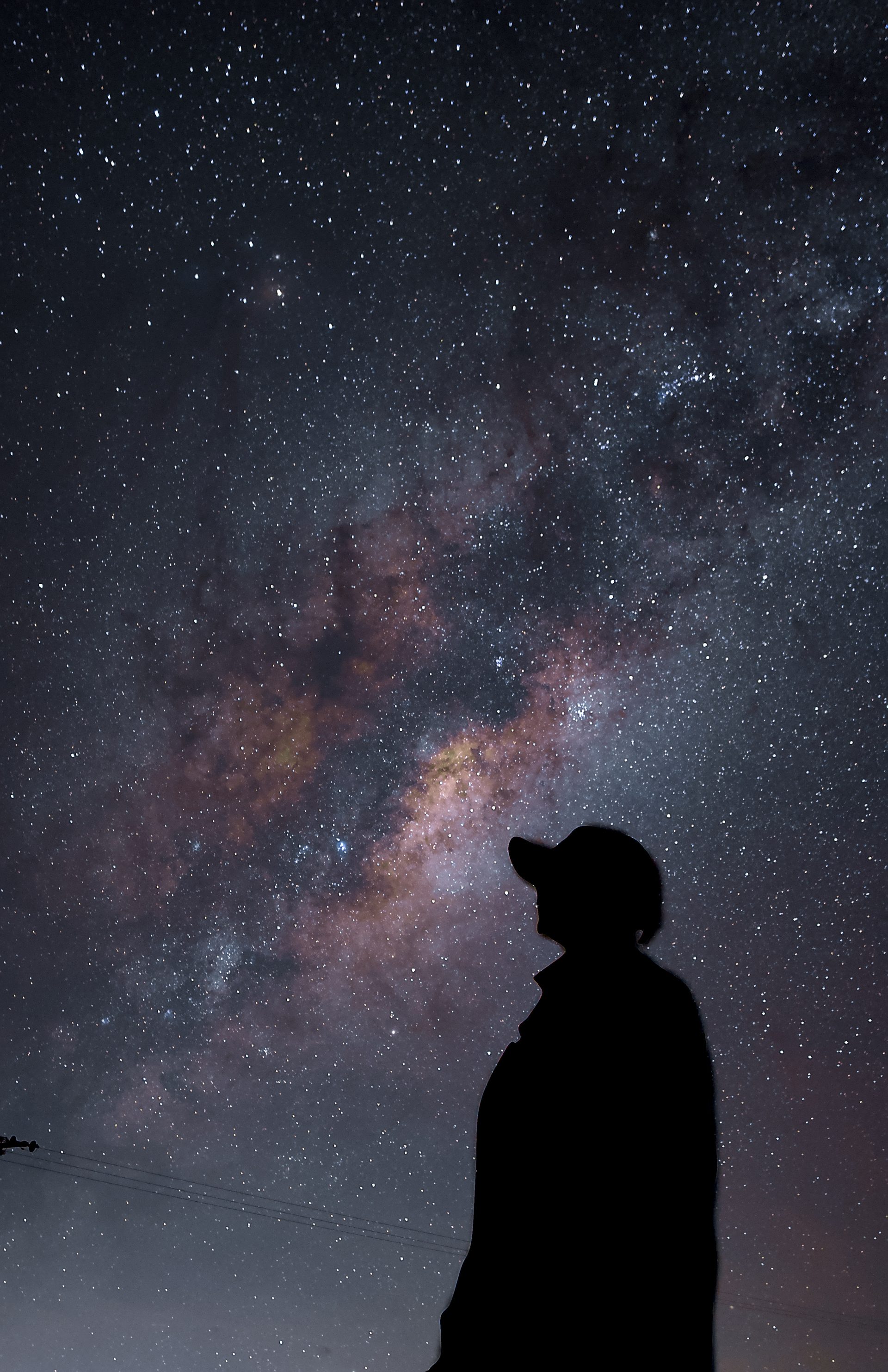 A person in a cap silhouetted against the Milky Way core, looking up, with power lines in the bottom corner.