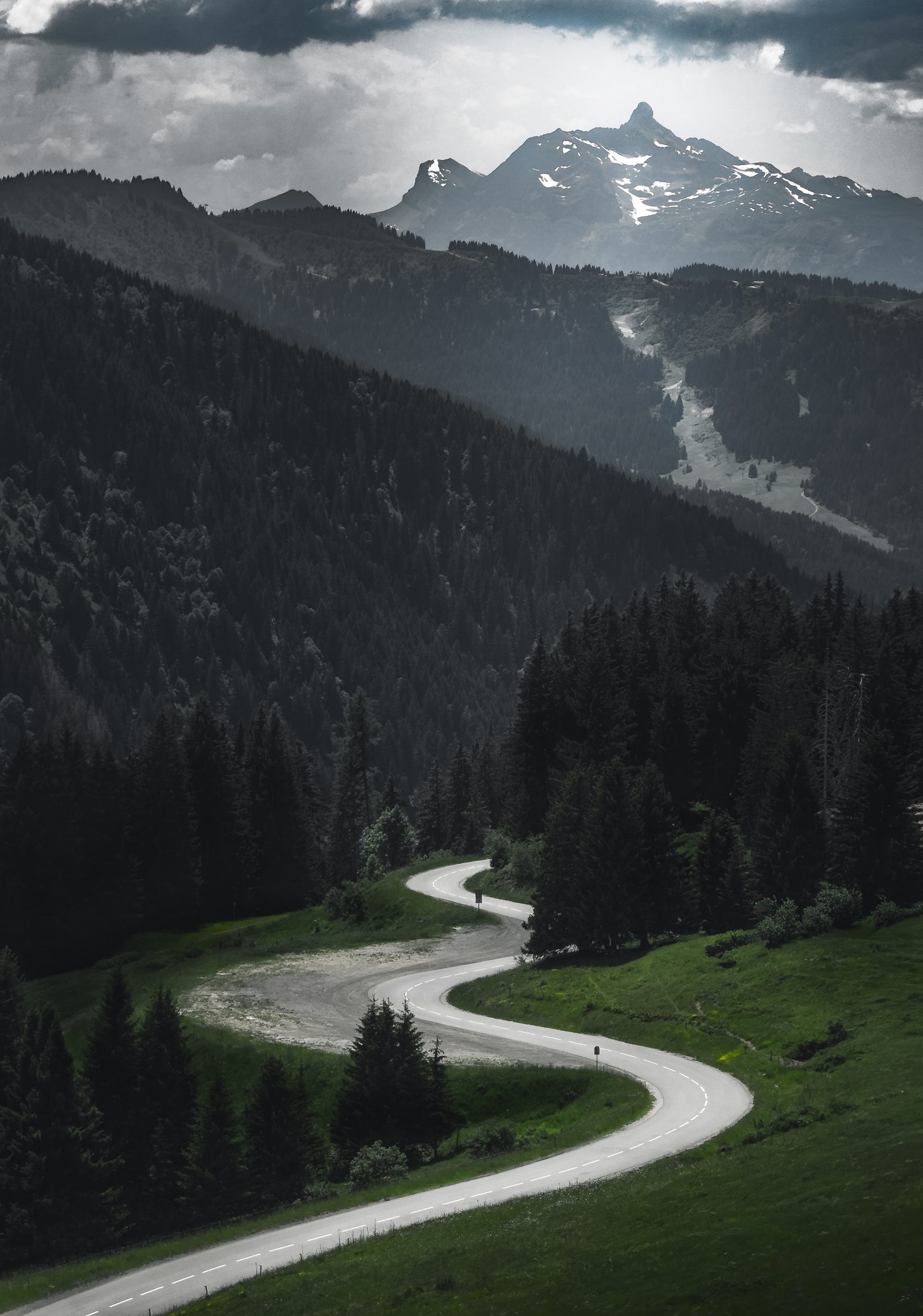 A road winding up a green hillside towards dark forested ridges, with a snow-streaked mountain peak far beyond under heavy cloud.