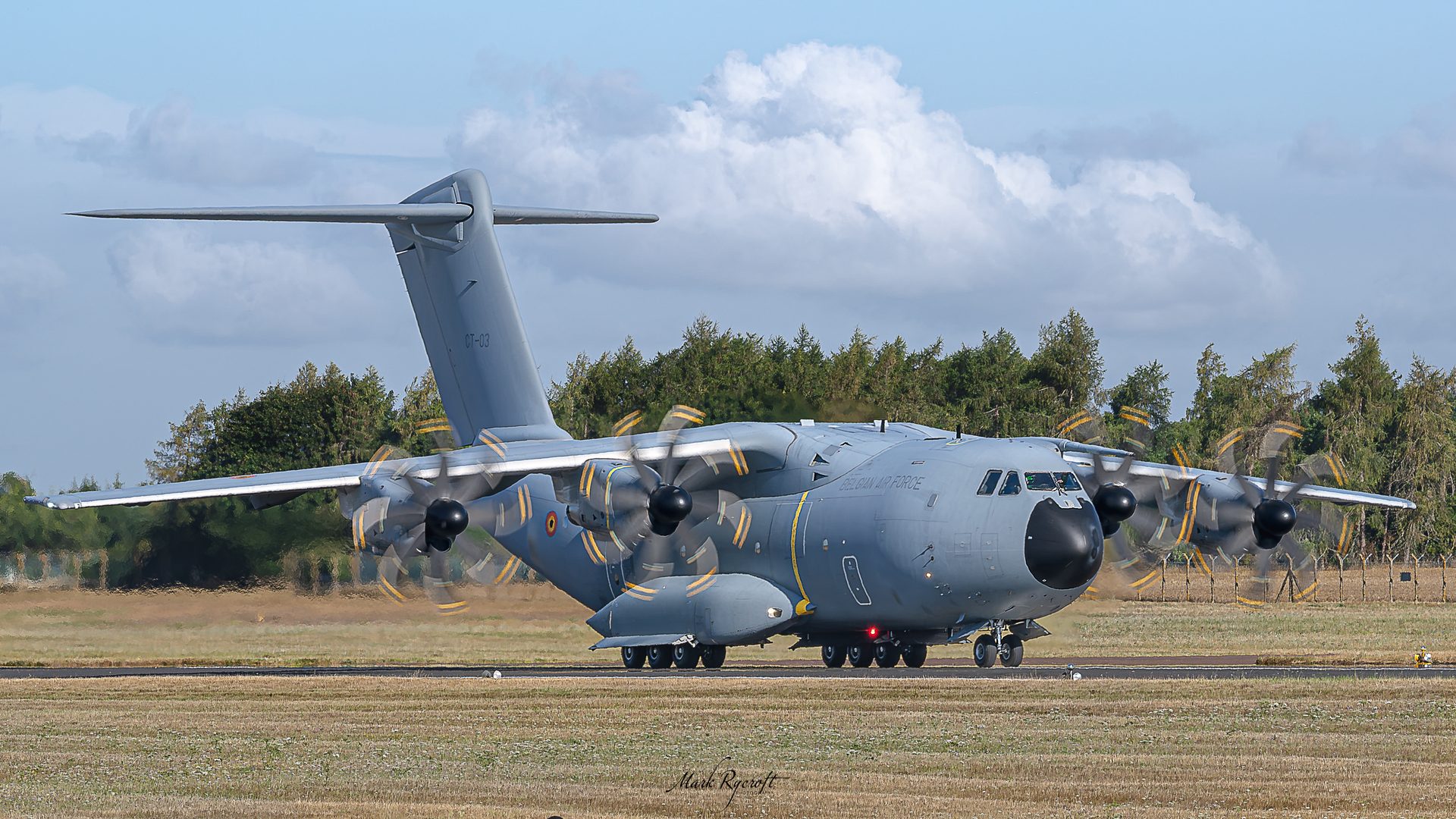 A grey Belgian Air Force A400M on the runway with all four propellers turning, trees and a cloudy sky behind.