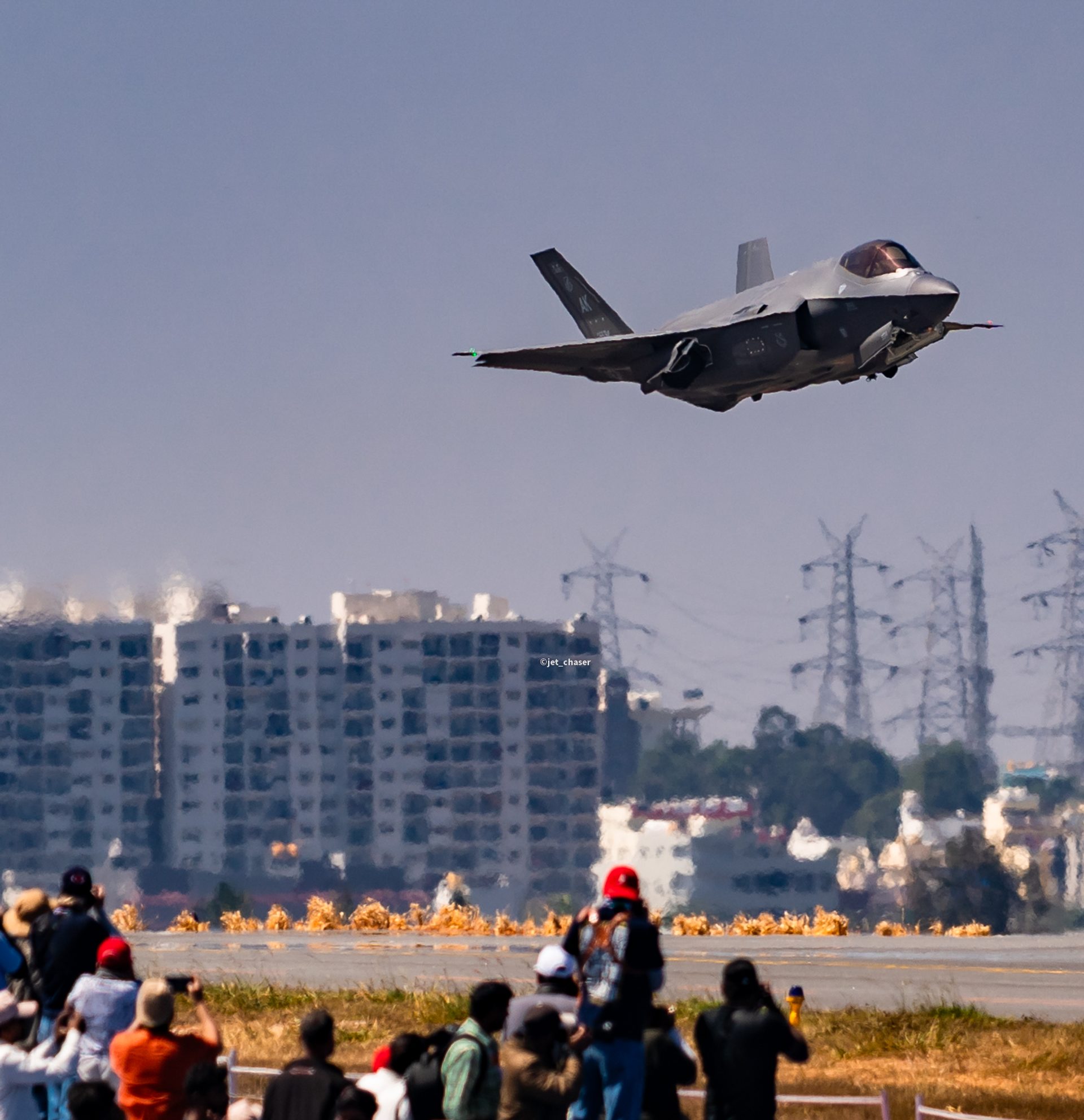An F-35 flying low over the runway at Aero India, with a crowd of spectators and photographers in the foreground and tower blocks behind