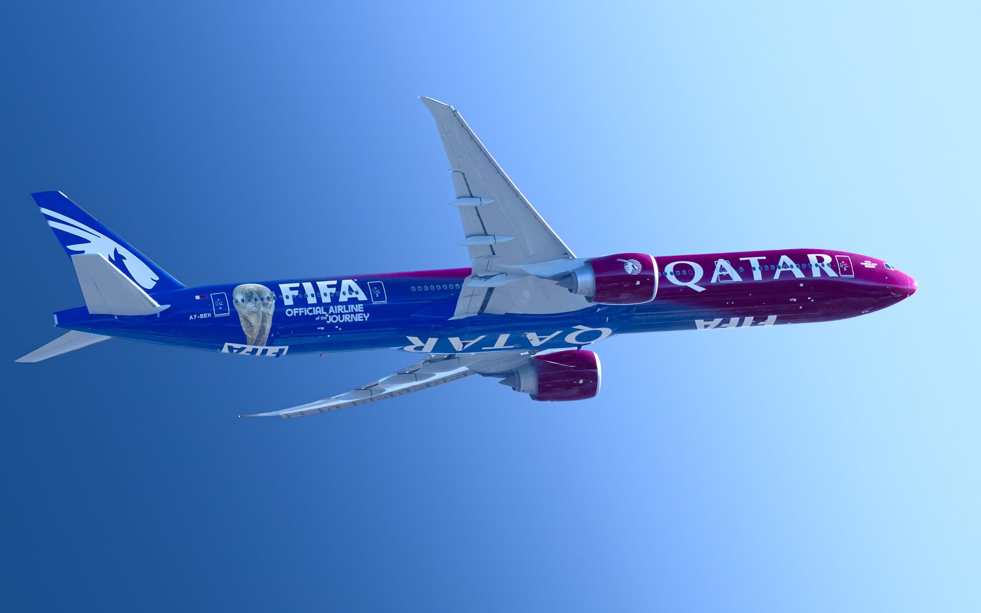 A Qatar Airways airliner in the blue and maroon FIFA livery, banking against a clear blue sky.